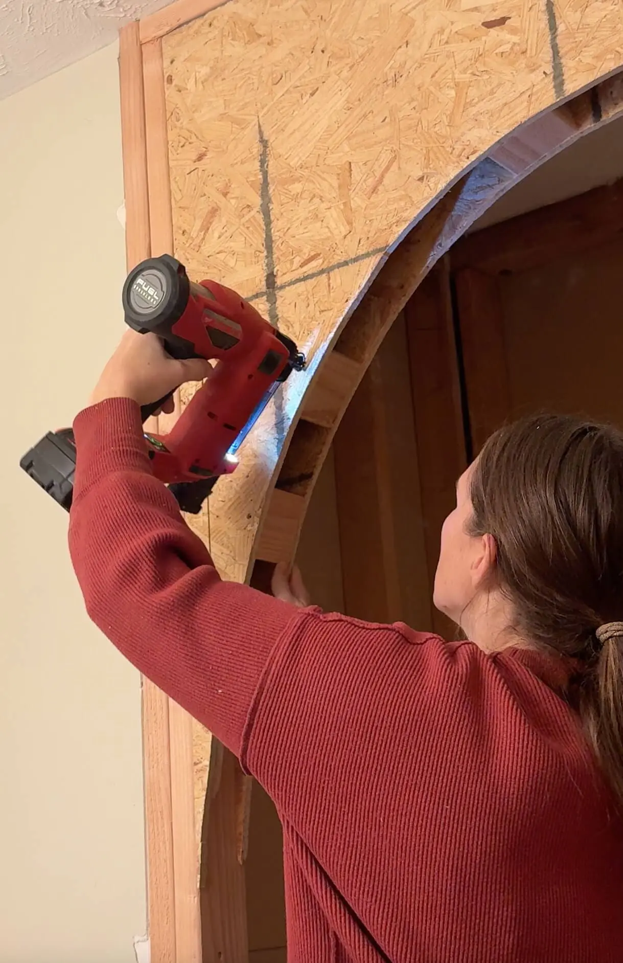 A close-up of a woman in a red sweater holding a red nail gun to fasten a curved OSB arch panel onto a wooden frame for an arched bathtub alcove. The detailed gridlines on the panel show the design planning for the remodel.